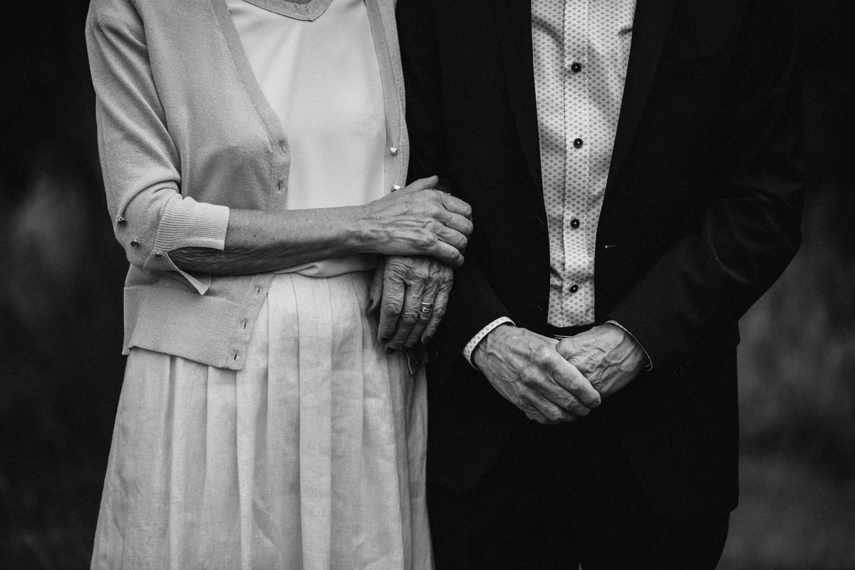 Casal de idosos de braços dados, ela com a mão sobre a dele, em foto preto e branco